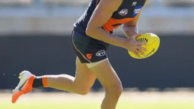 A professional Australian rules footballer sits on the bench during a match, visibly in discomfort.