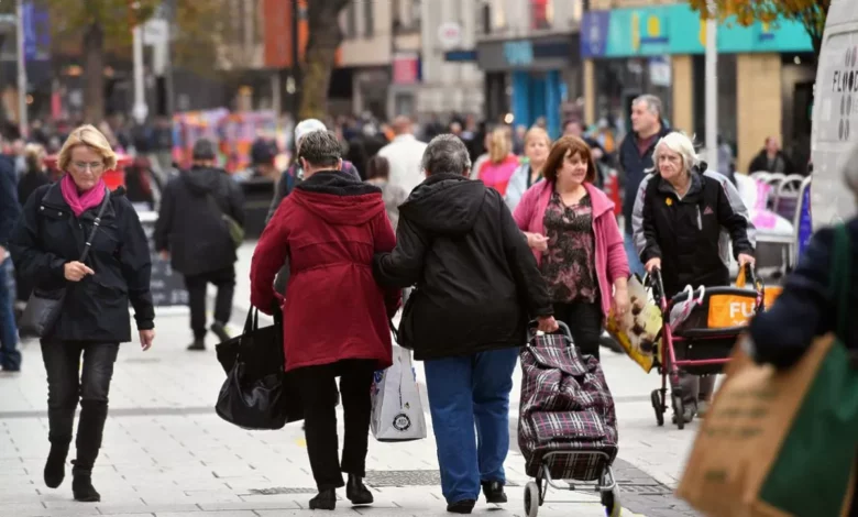 Shoppers walking past a row of shops with sale signs in a Welsh high street.