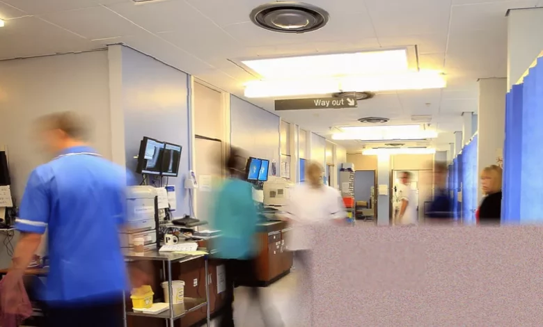 Crowded hospital emergency department waiting area with patients on chairs and staff at a counter in England.