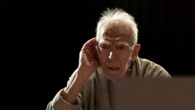 An elderly actor on a dimly lit stage with a reel-to-reel tape recorder