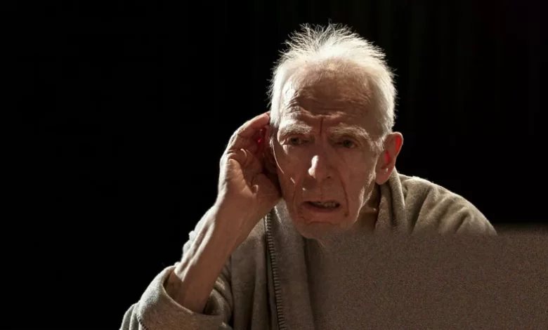 An elderly actor on a dimly lit stage with a reel-to-reel tape recorder